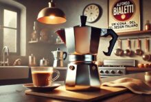 Caffettiera Moka Bialetti in acciaio su un piano di cucina, accanto a una tazza di caffè e alcuni biscotti disegnati artisticamente, con dettagli vintage sullo sfondo.